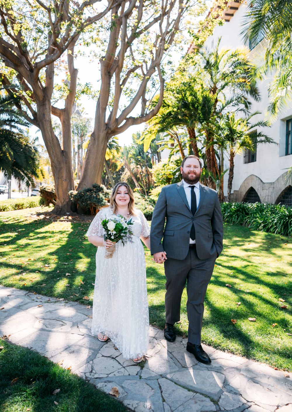 Wedding couple poses in formal attire on stone path surrounded by tropical garden and architecture.