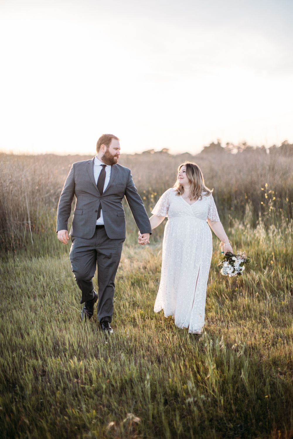 Couple walks hand in hand through golden field at sunset in wedding attire.