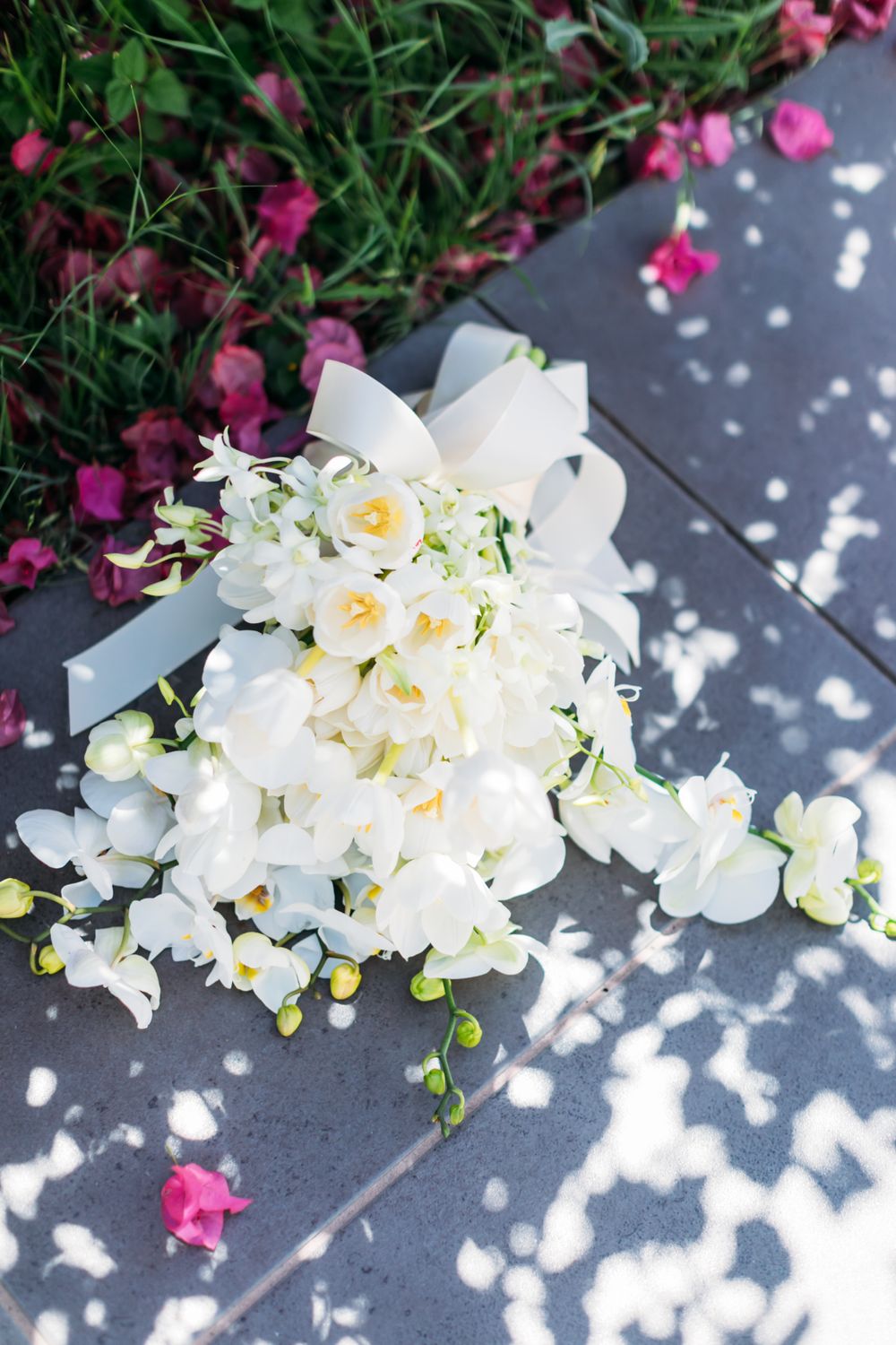 Elegant white orchid and rose bridal bouquet with silver ribbon resting on concrete in dappled sunlight with pink flowers.