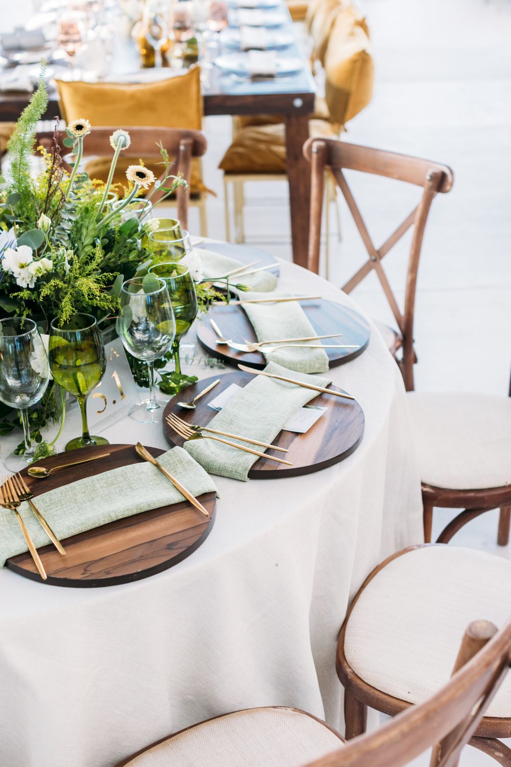 Elegant table setting with wooden chargers, white tablecloth, cross-back chairs and fresh greenery centerpiece at wedding reception.