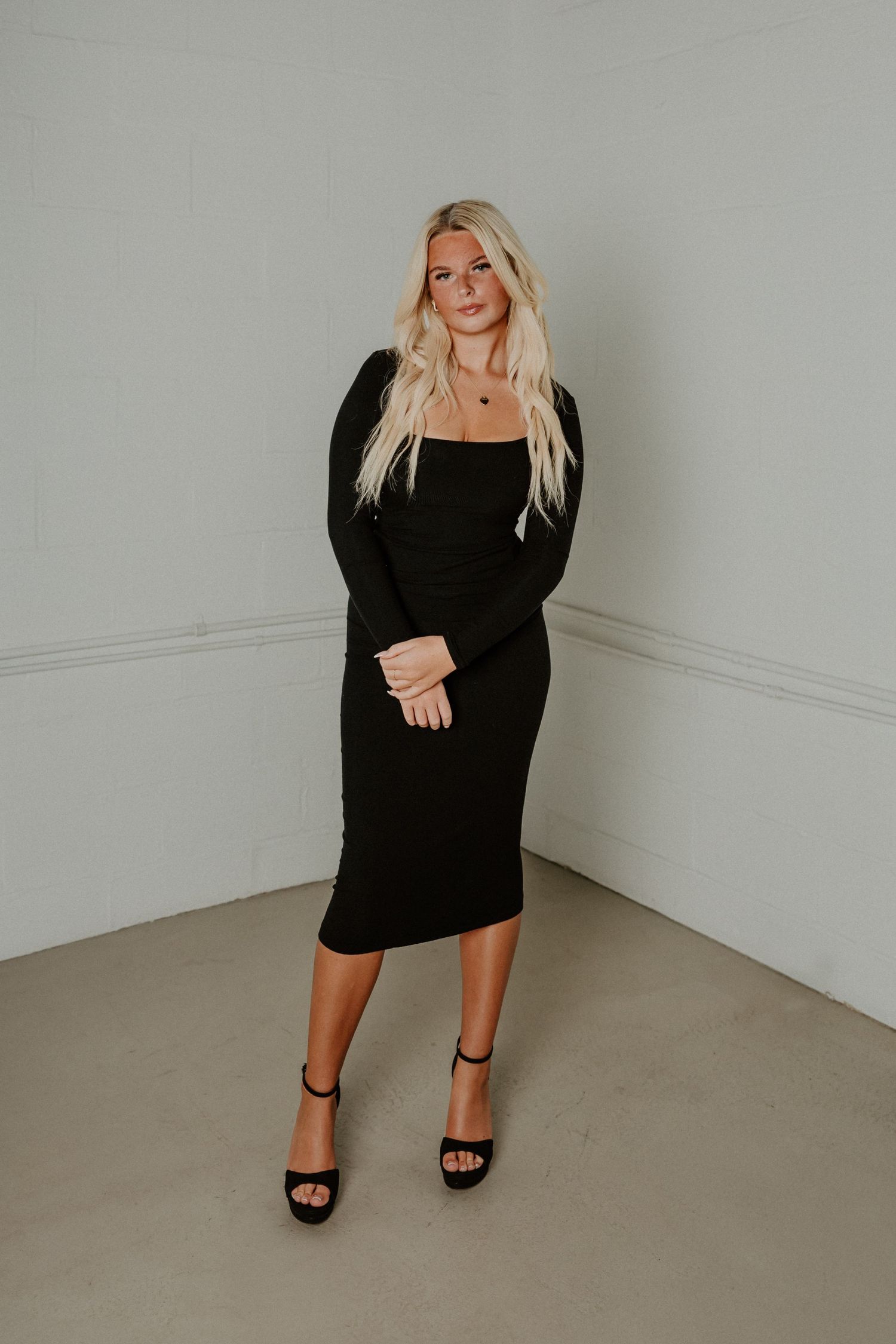 A woman in a black long-sleeve midi dress poses against a white wall wearing platform heels.