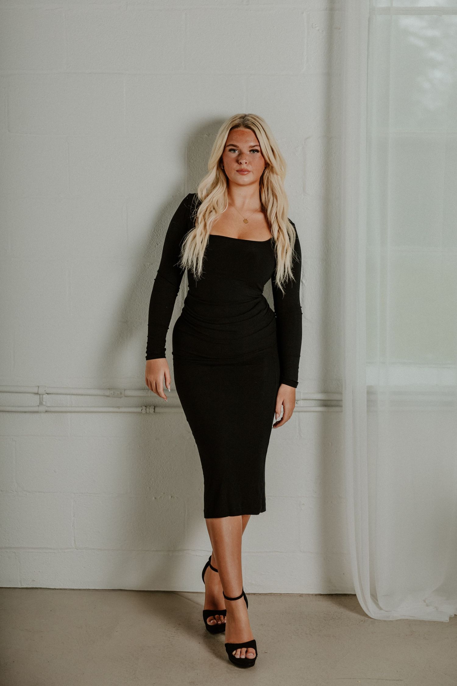 Elegant long black dress with long sleeves and ankle strap heels in minimal studio setting.