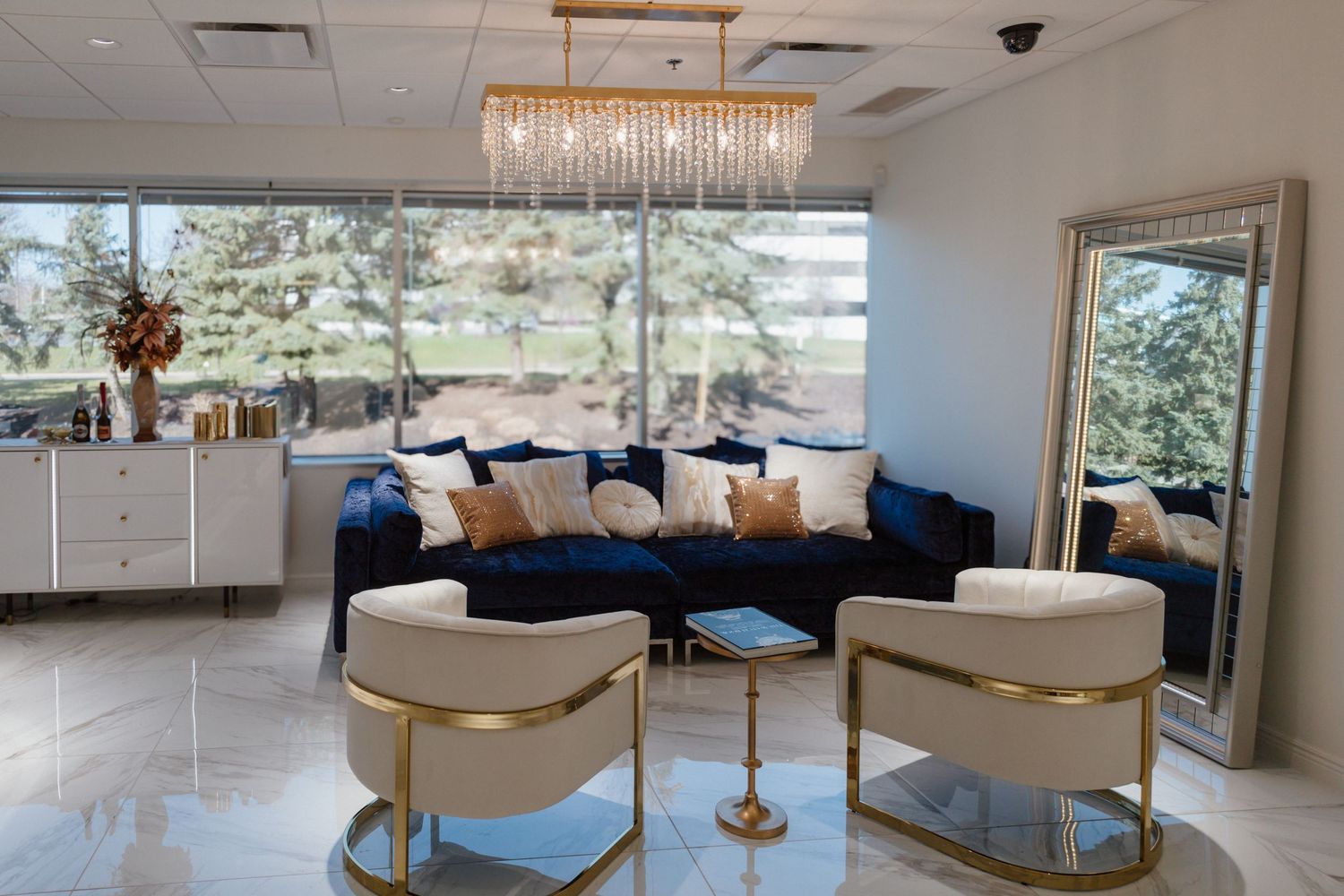 Modern luxury waiting area with blue velvet seating, gold accents, and a crystal chandelier in a bright space.