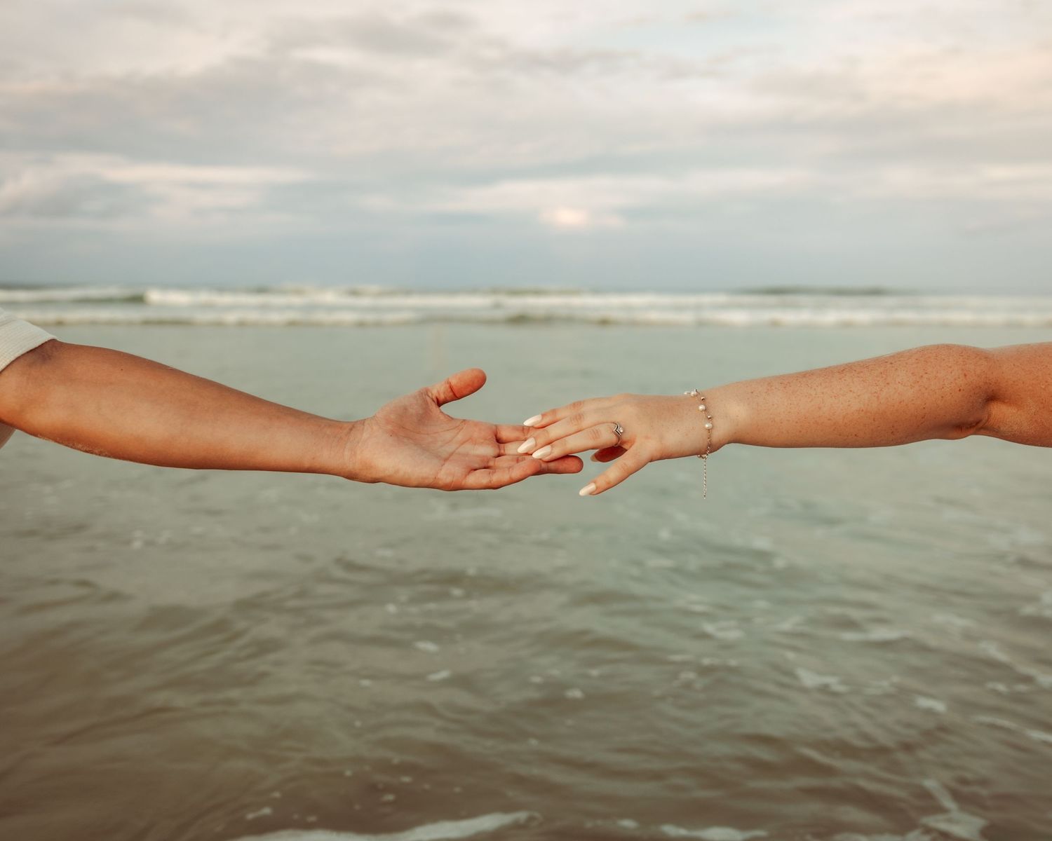 Close-up of couple's hands with tattoos and jewelry holding each other against sunset backdrop.