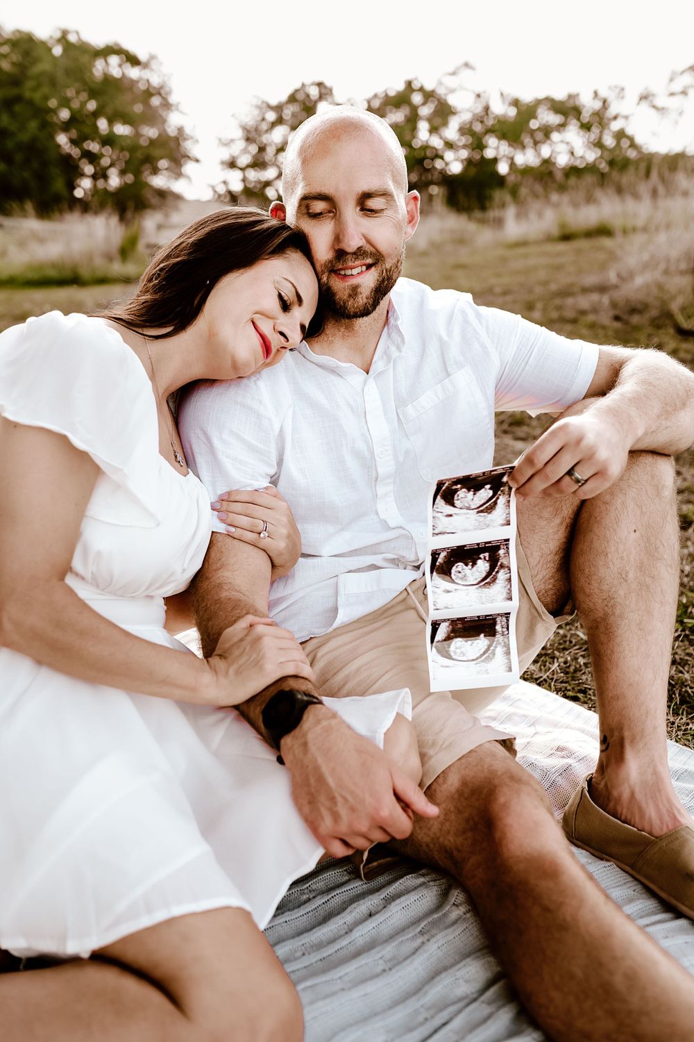 A couple sits on blanket outdoors holding ultrasound photos.
