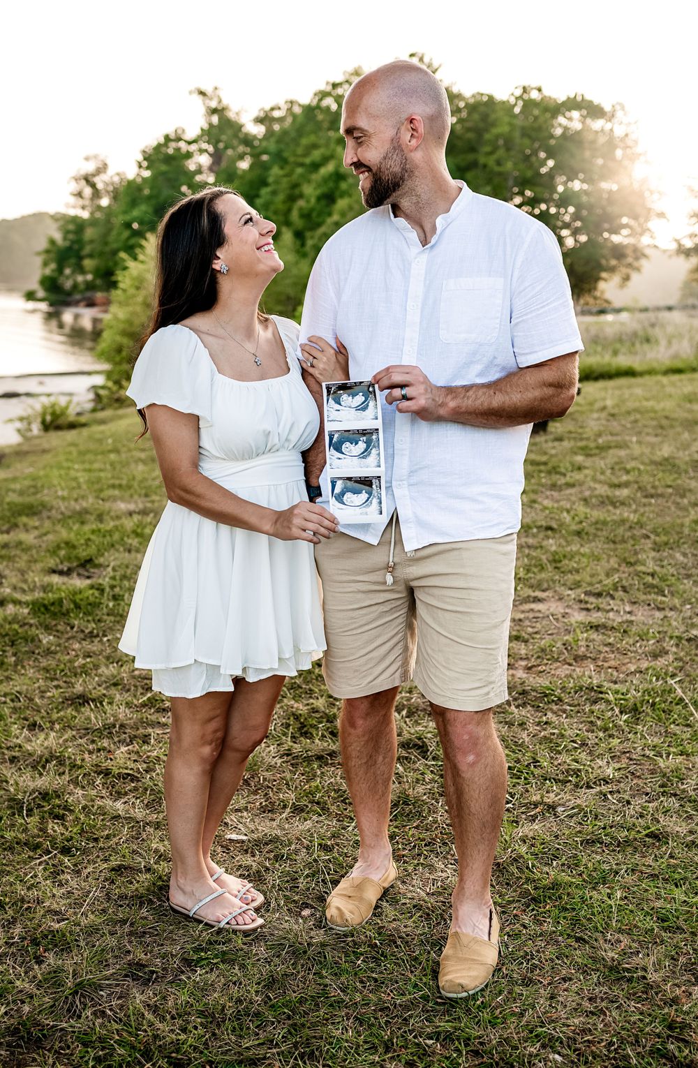 A couple in white outfits holding ultrasound photos stands in a field at sunset.