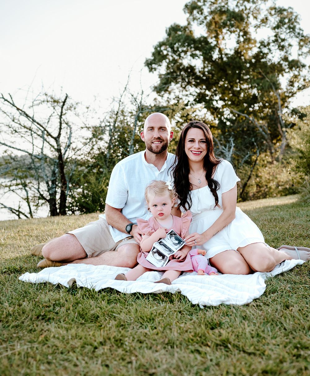 Family of three sitting on white blanket outdoors during golden hour photoshoot.
