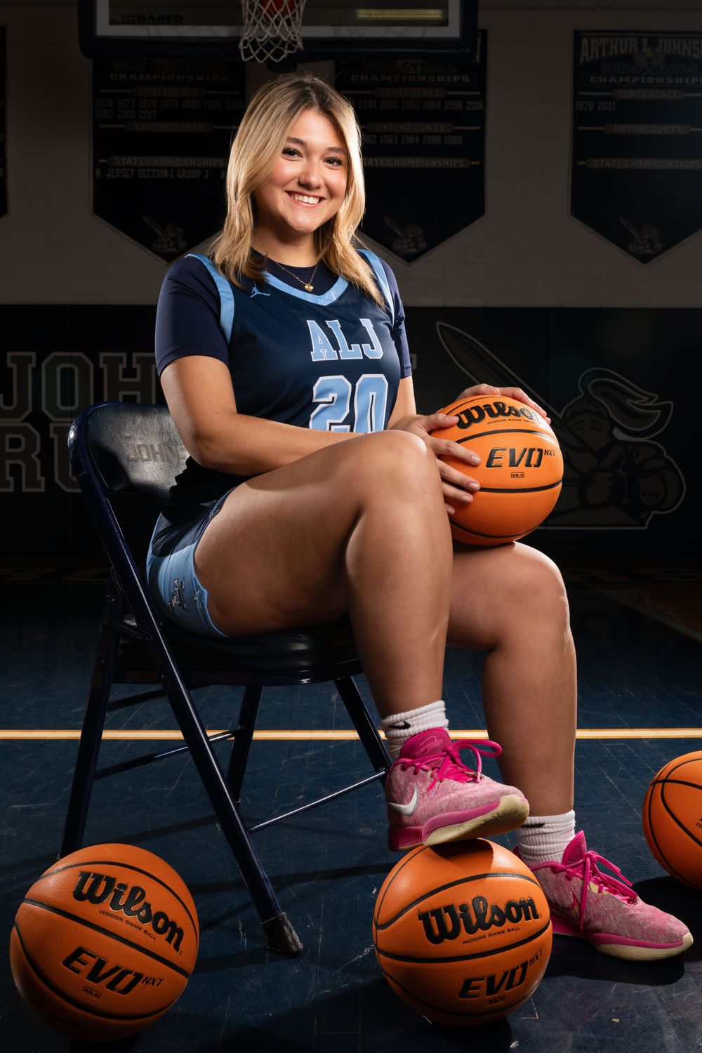 Basketball player poses on chair with Wilson basketballs in school gym setting.
