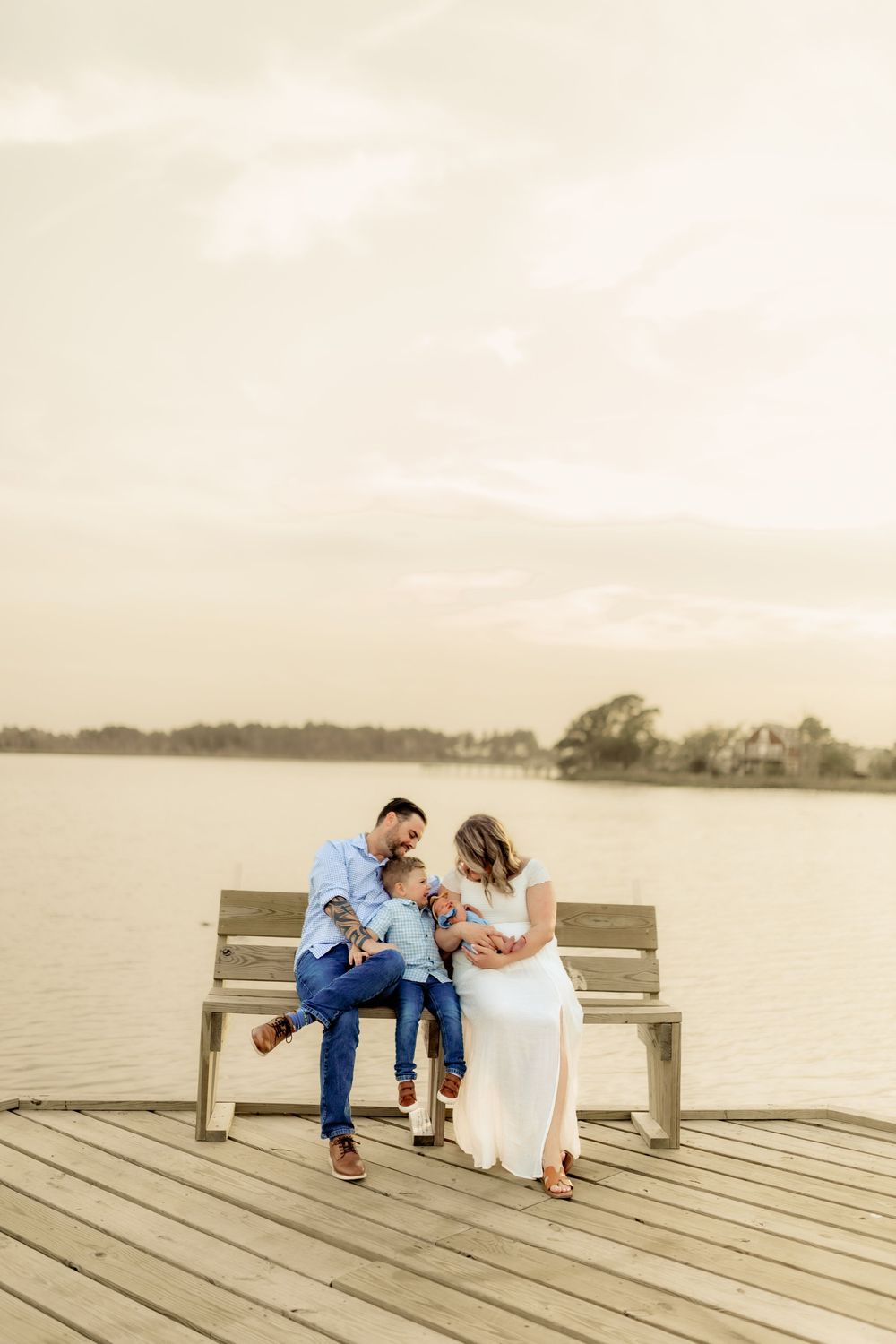 Family of three sitting on a wooden dock bench by the water, sharing a tender moment at golden hour.