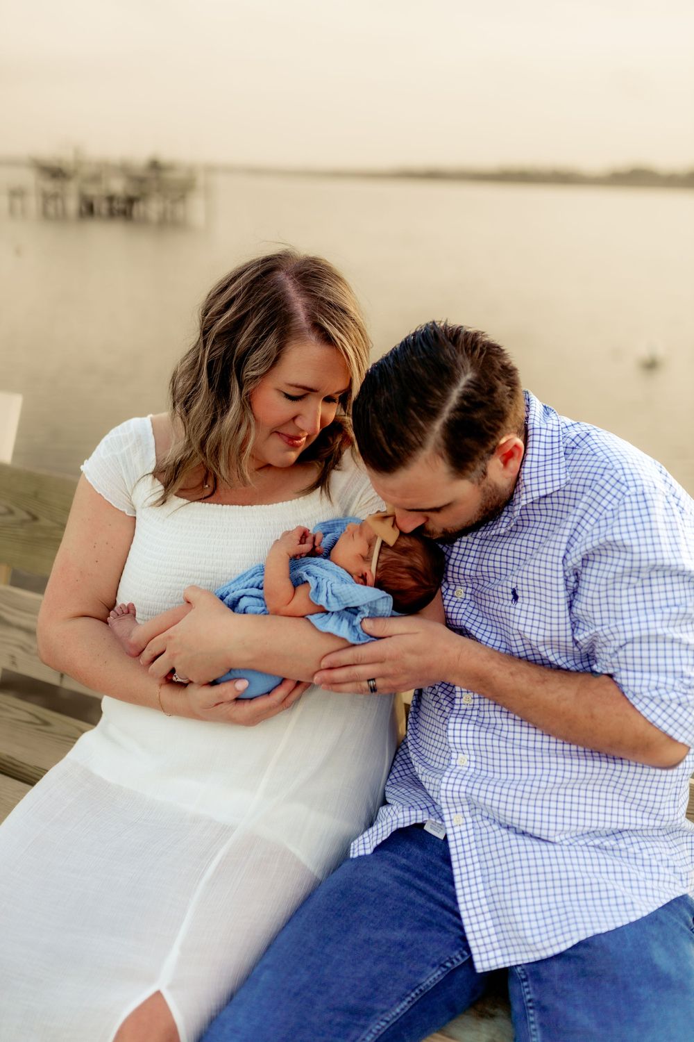 A couple sits outdoors near water, lovingly cradling their newborn baby wrapped in a blue blanket.
