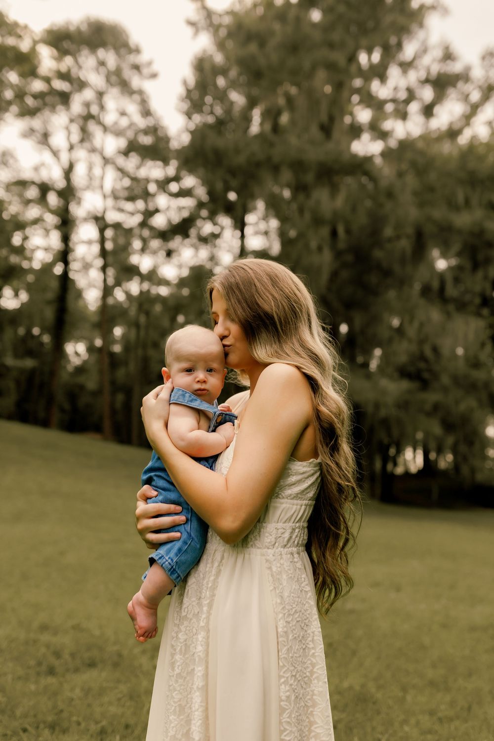 A mother in a white lace dress holds her baby outdoors on a green lawn surrounded by tall trees.