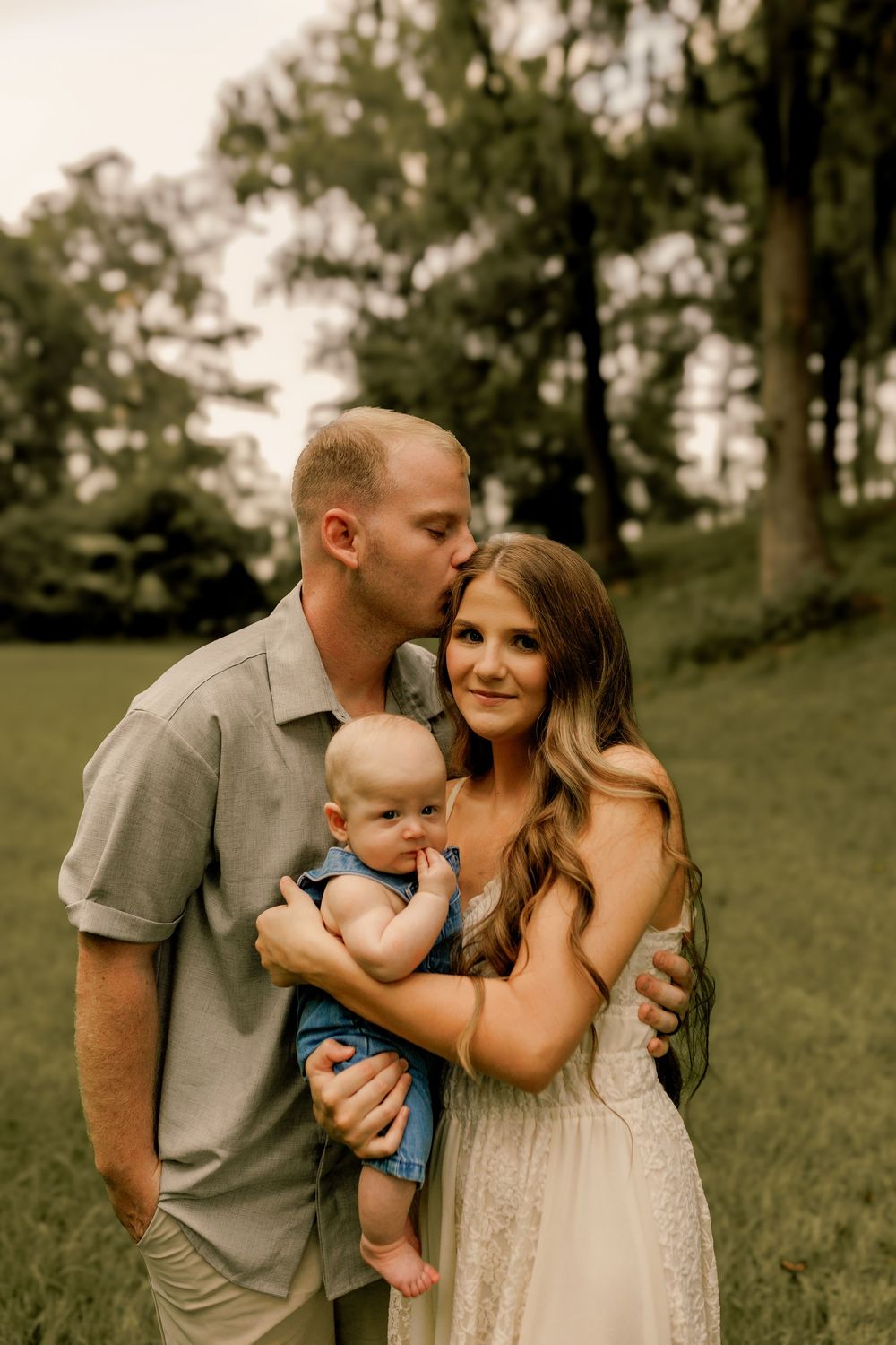 A man kisses a woman on the cheek as she holds their baby outdoors in a lush green park setting.
