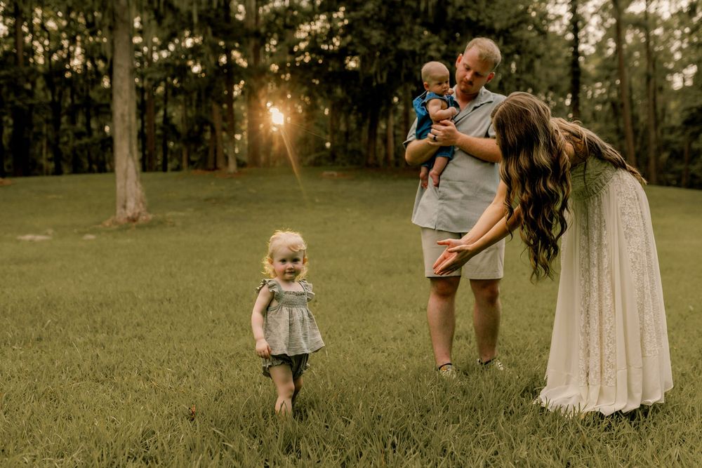 Family of four enjoying golden hour outdoors, parents smiling at baby while toddler runs across lush green grass.