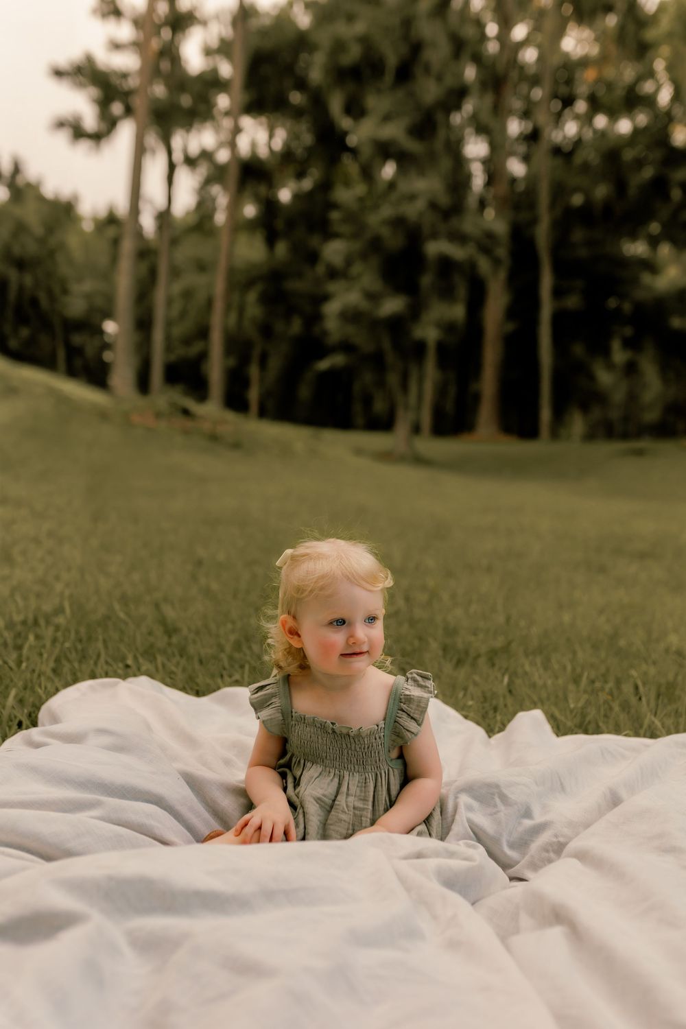 Toddler girl in olive dress sitting on white blanket in lush green park at golden hour.