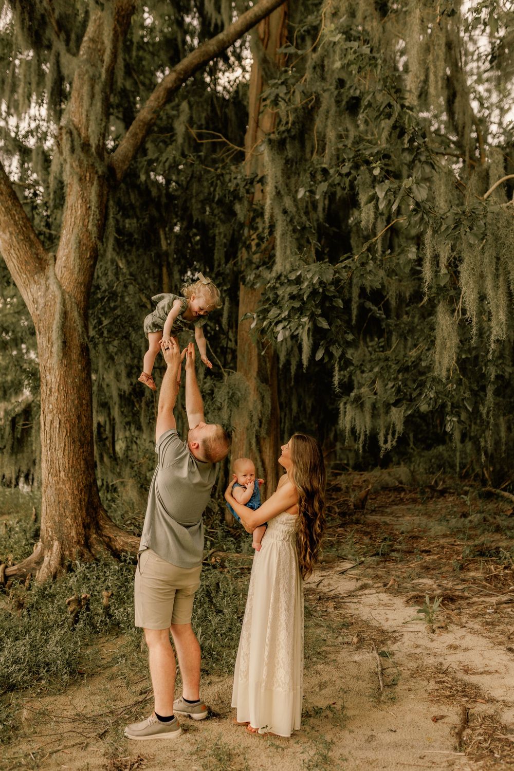A family poses outdoors under moss-draped trees, father lifting toddler while mother holds infant in a white dress.