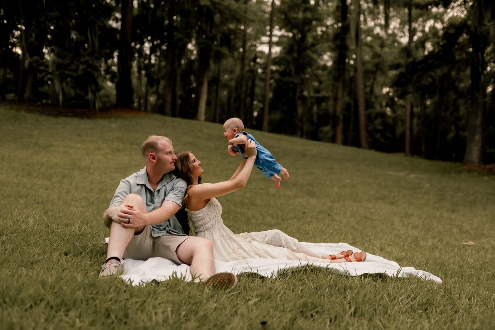 Family sitting on blanket in park, parents lifting baby in blue outfit, surrounded by lush green trees.