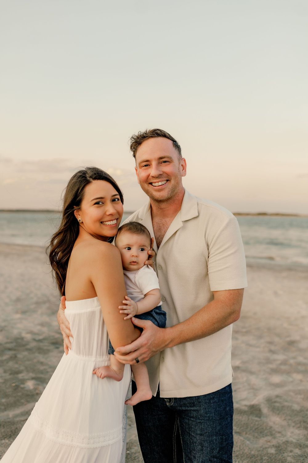Happy young family with baby posing on a sandy beach at sunset, wearing white and neutral tones.