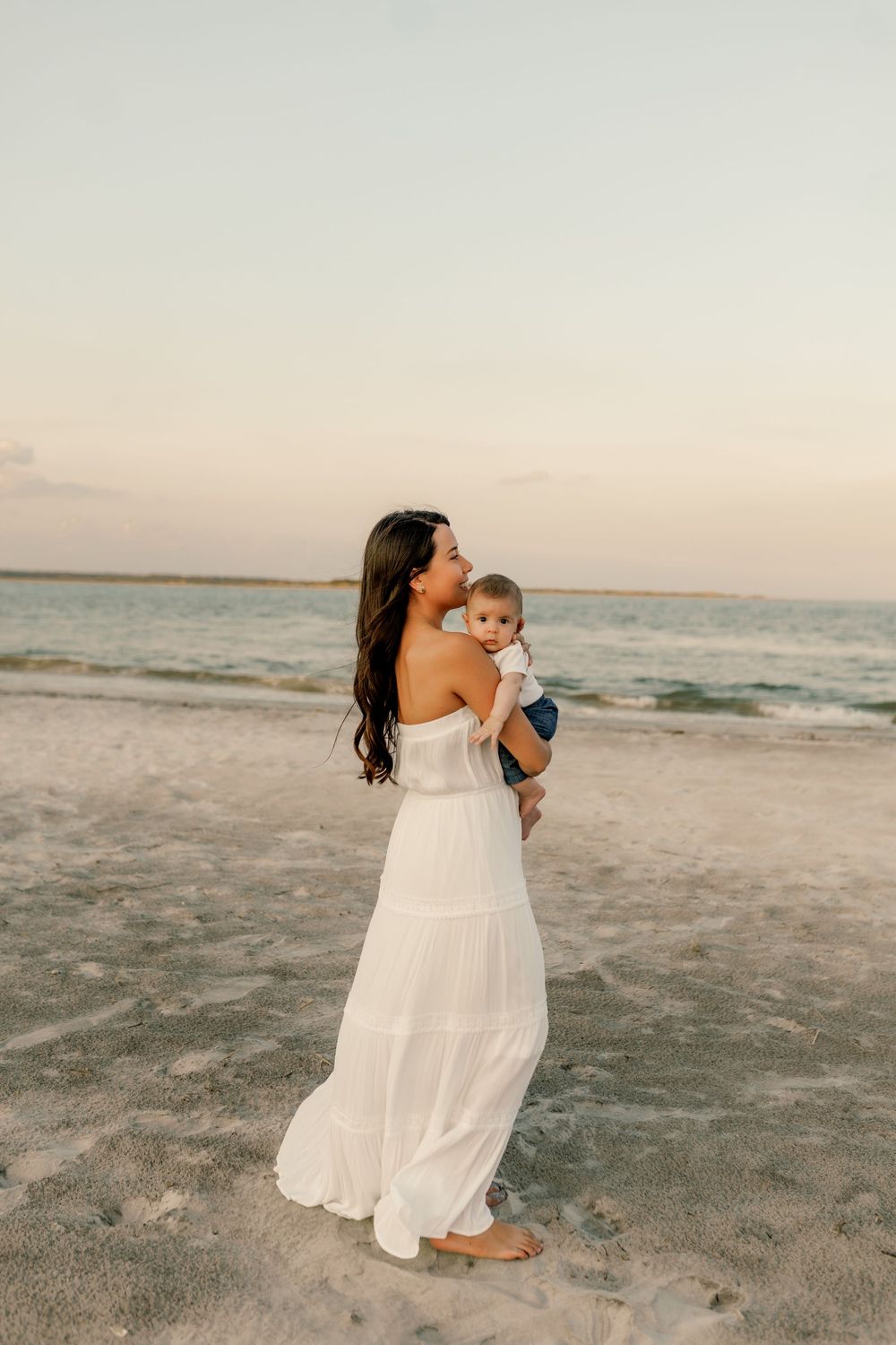A woman in a white maxi dress holds her baby on a sandy beach at sunset.