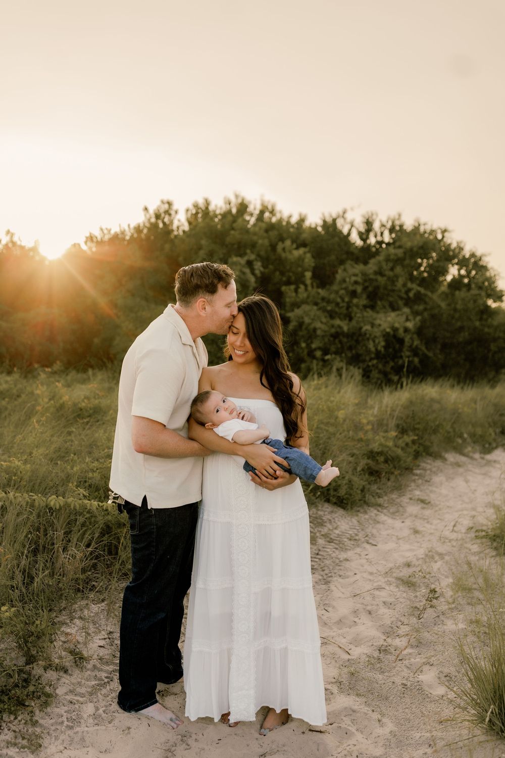 A couple shares a tender moment at sunset on a sandy path, lovingly holding their newborn baby outdoors.