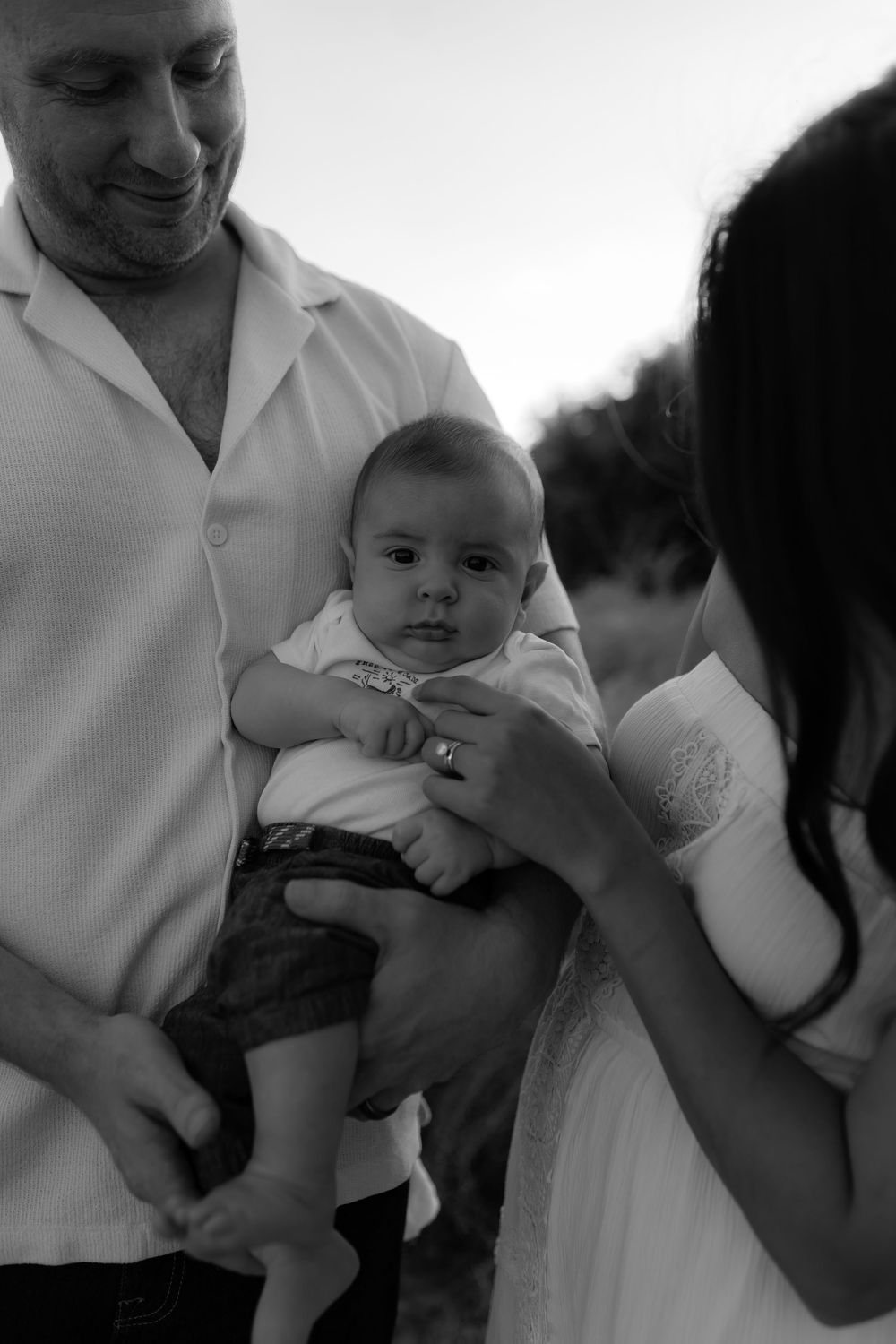 Black and white photo of a smiling man holding a baby while a woman in white dress looks on tenderly.