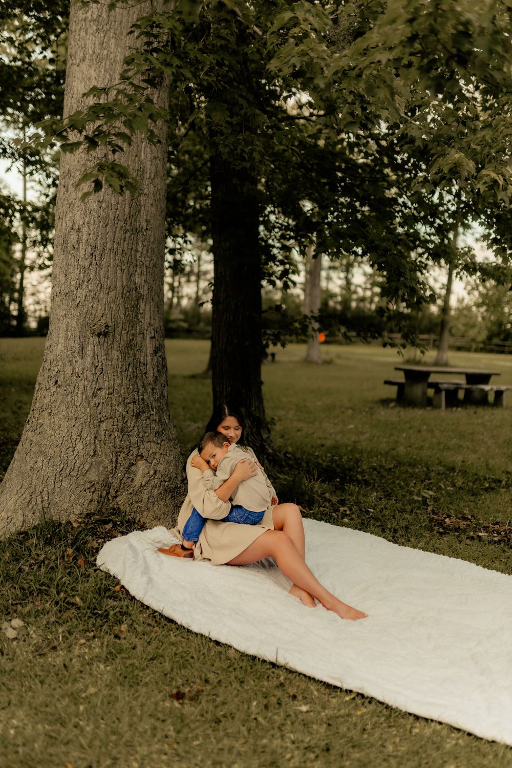 A woman sits barefoot on a white blanket under a large tree in a park, cradling a baby in her arms.