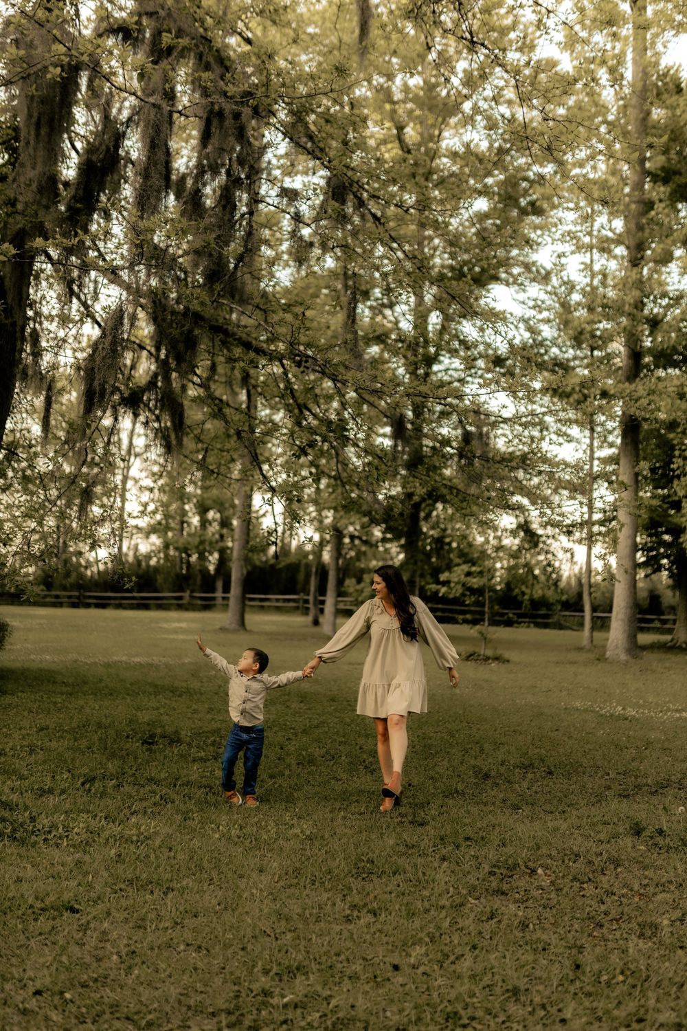 A woman in a white dress holds hands with a young child in a lush green park surrounded by tall trees.