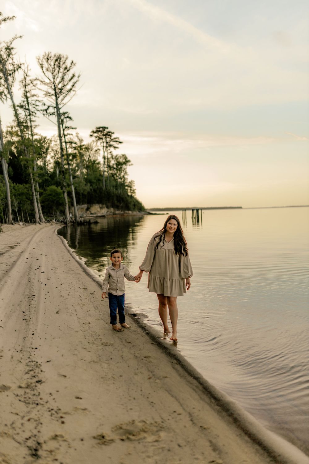 A woman and young child hold hands walking along a sandy beach shoreline at golden hour sunset.