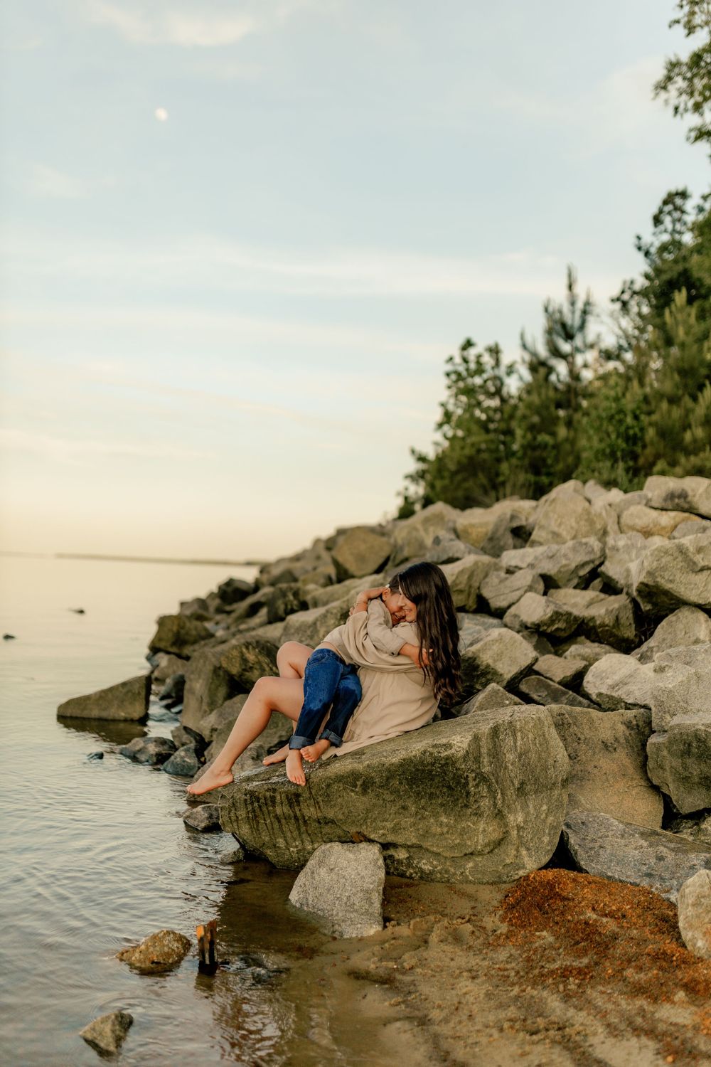A mother and young child sit together on rocky shoreline at sunset, sharing a tender embrace by calm waters.