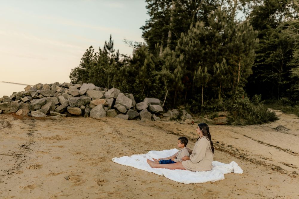 A woman and child sit on a white blanket on a sandy beach surrounded by rocks and lush green trees at sunset.