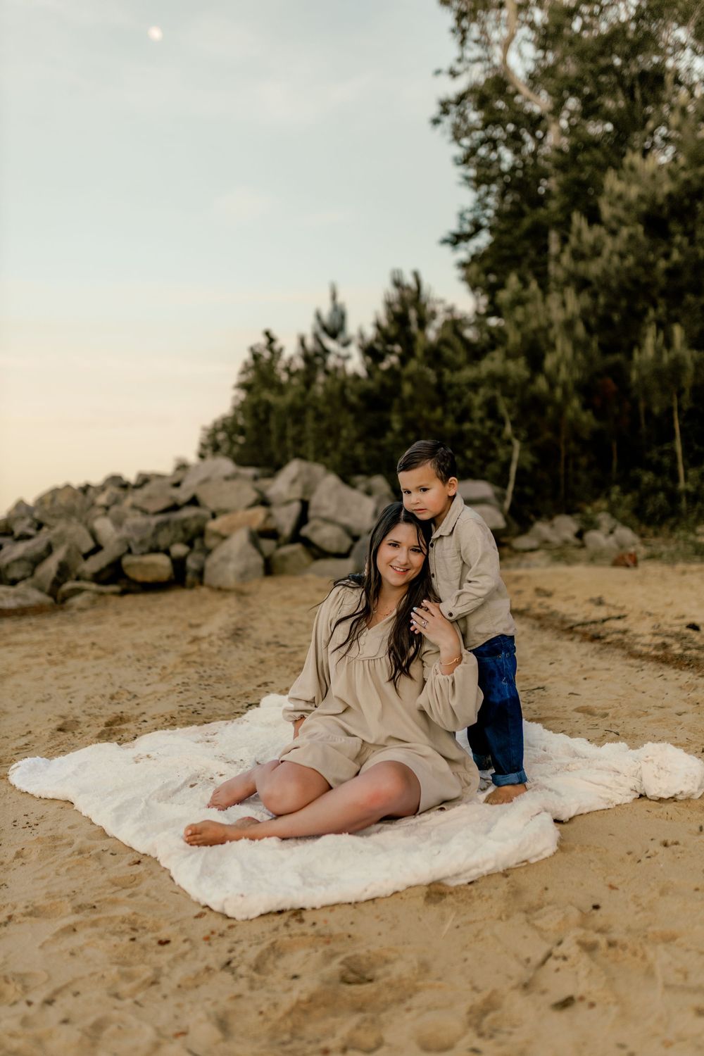 A pregnant mother sits on a white blanket on sandy beach at dusk, smiling as her young son stands behind her.