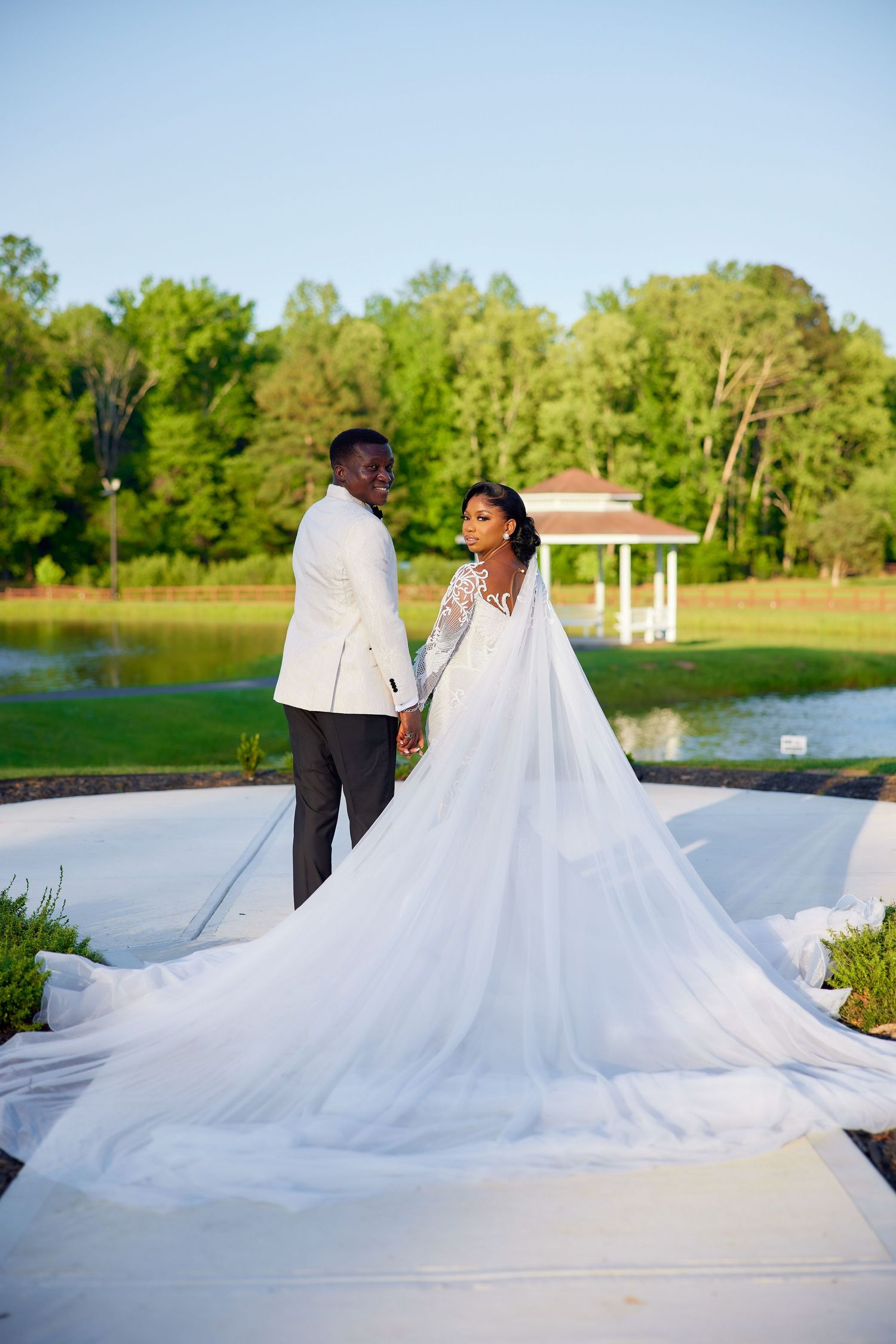 A stunning wedding couple poses on a white platform near a lake with their flowing white dress spread beautifully across the deck.