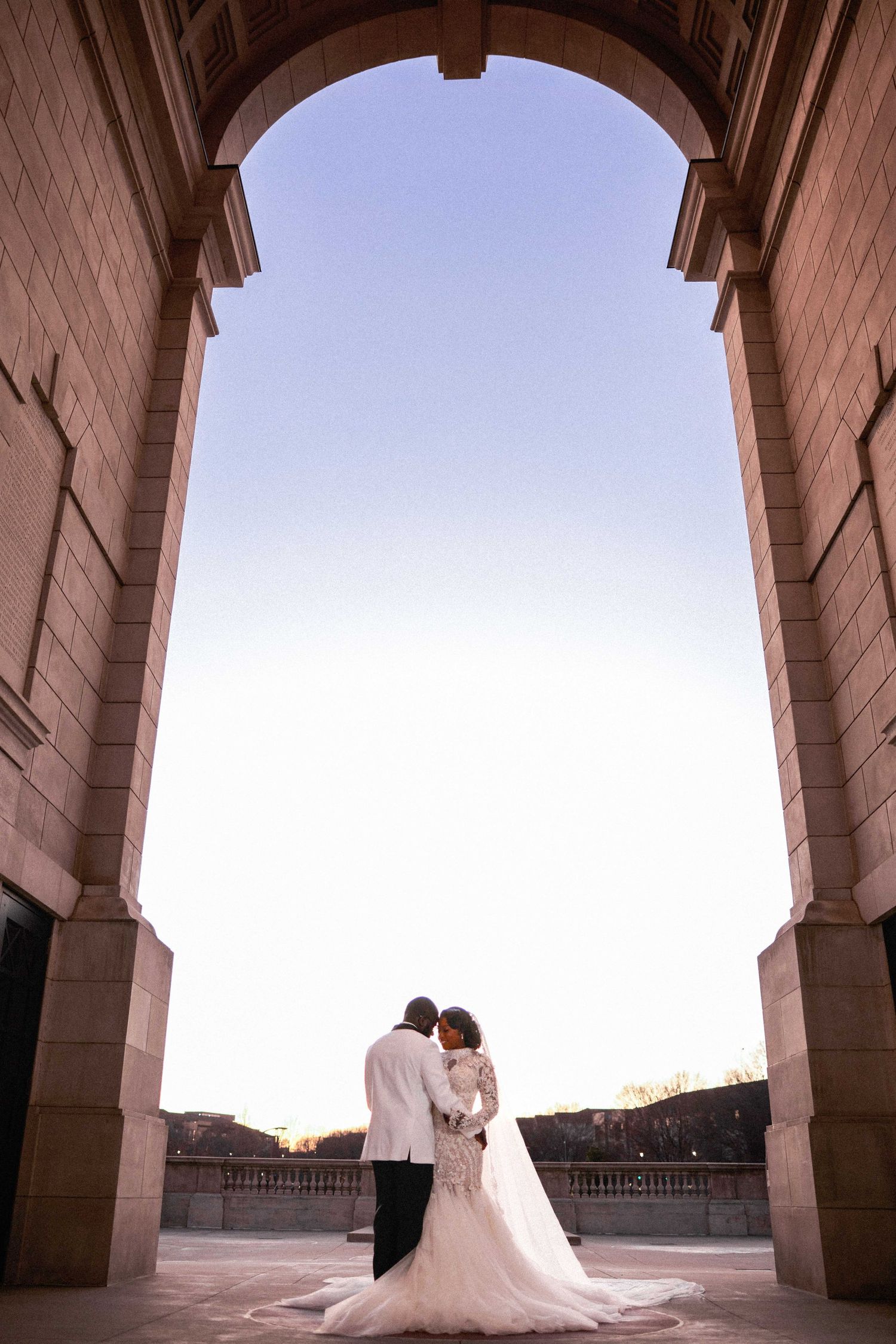 A couple in wedding attire stands beneath a grand stone archway during sunset, creating a romantic silhouette.