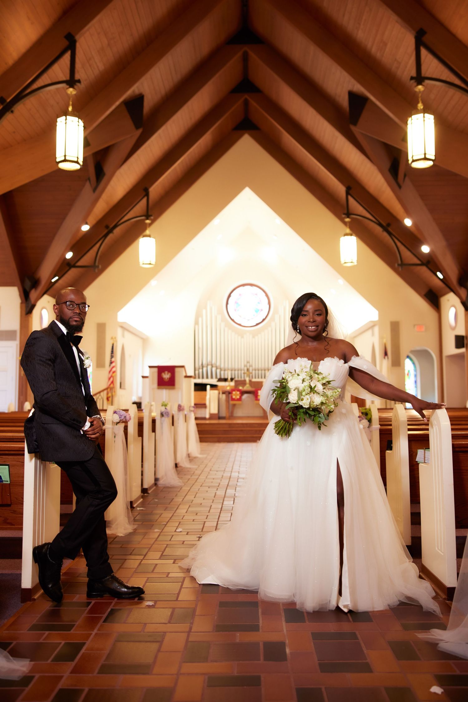 A wedding couple poses in the aisle of a beautiful church with wooden beams and hanging pendant lights.