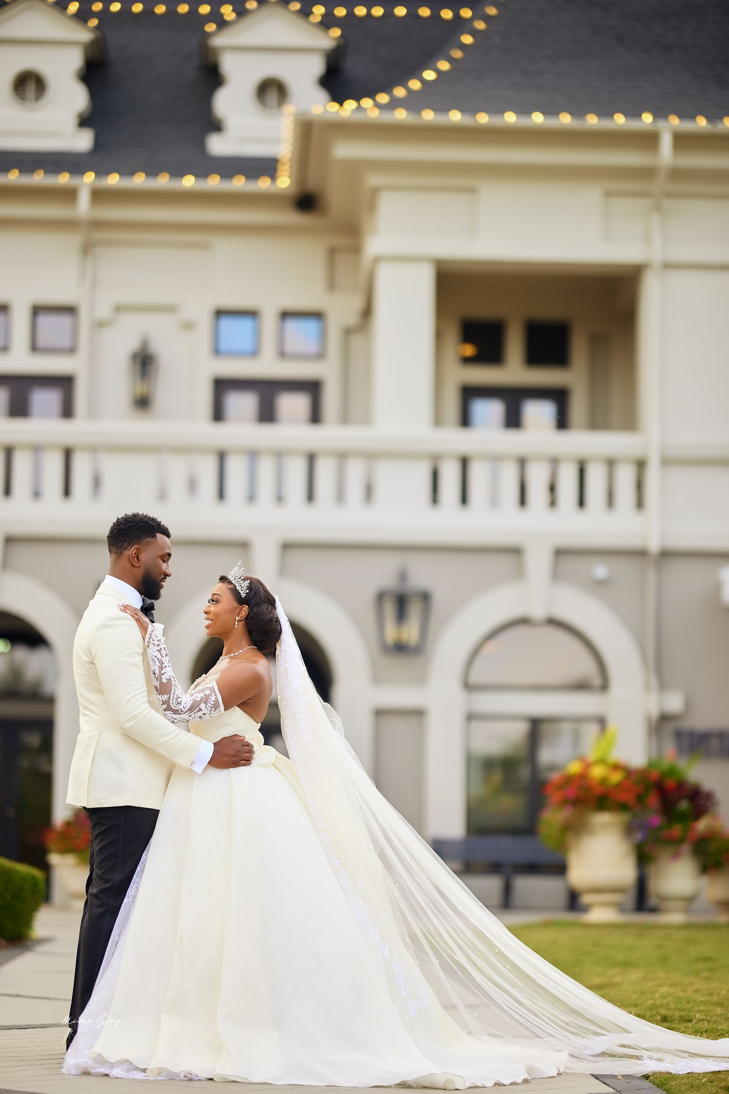 A joyful wedding couple embraces outside an elegant white mansion with string lights and ornate architectural details.