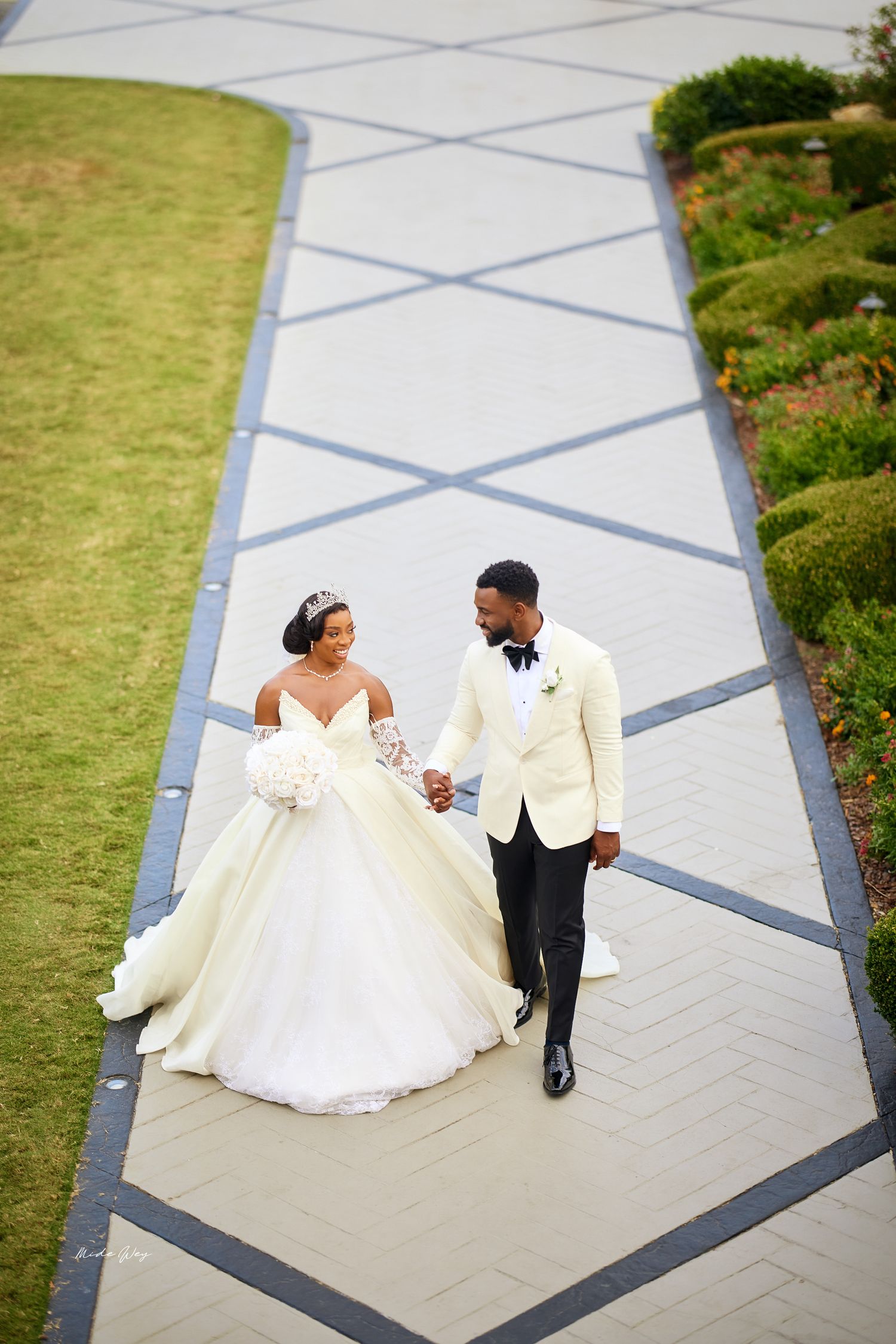 A couple in wedding attire poses on a geometric patterned walkway surrounded by green landscaping.