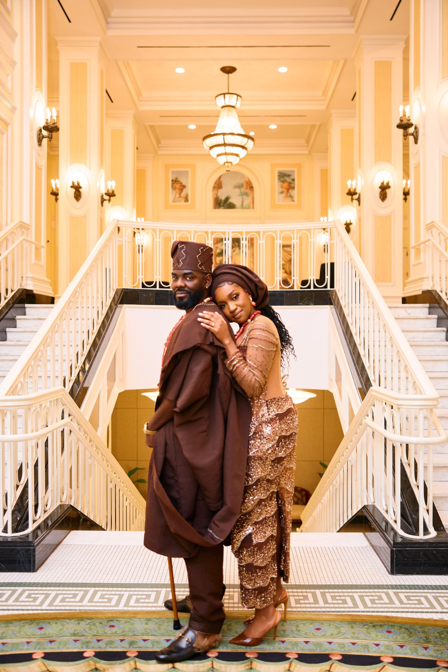 A couple embraces in an elegant hotel lobby with grand white staircases, ornate chandeliers and classic architectural details.