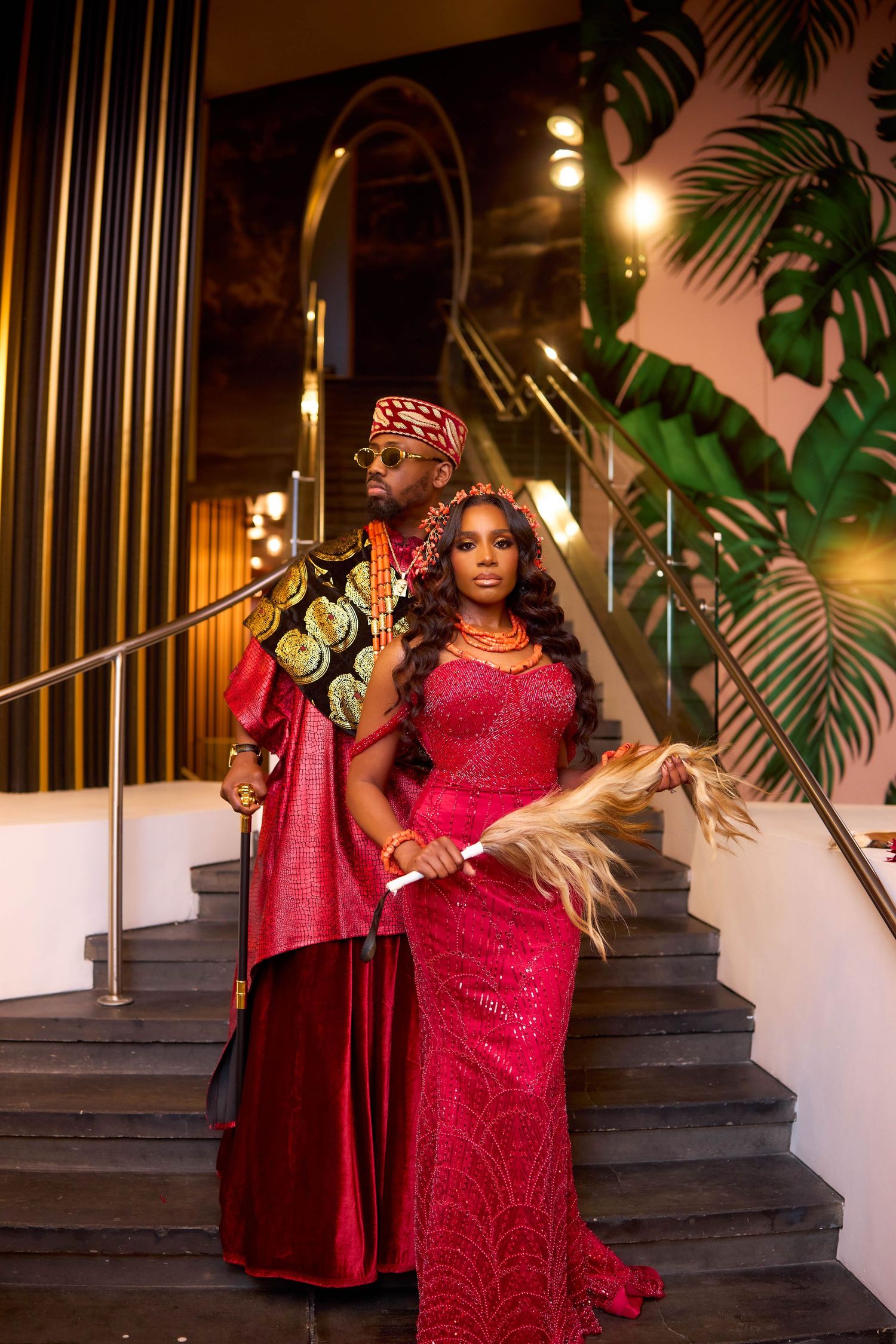 Two people in elegant red attire pose on a staircase with tropical decor and warm lighting in the background.