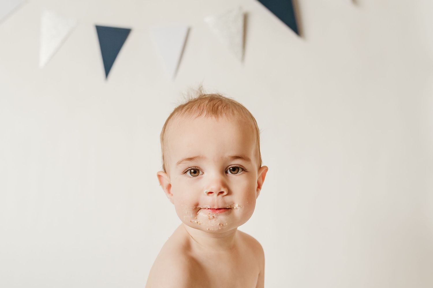 baby boy smiling at the camera with cake on his face and a birthday banner in the backdrop