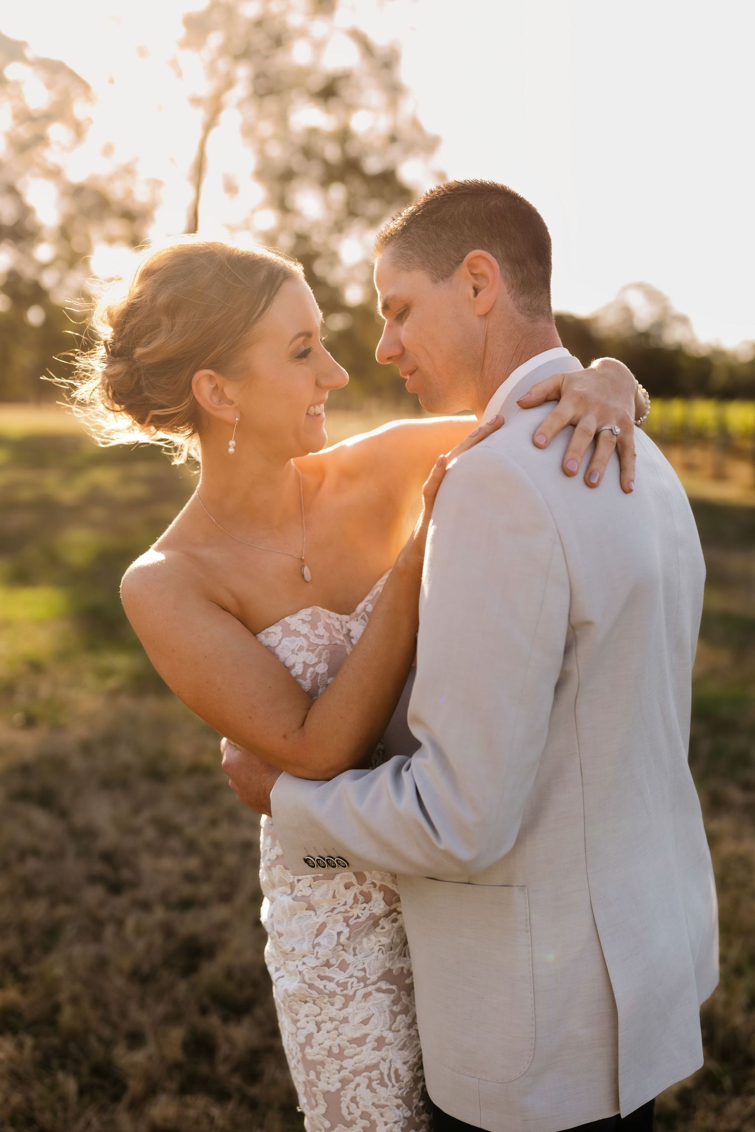 A couple dressed in wedding attire embrace during golden hour with sunlit trees in the background at sunset. Narkoojee Winery. Gippsland.