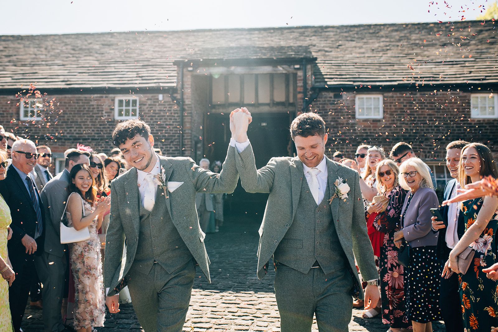 Wedding party celebrates with confetti and joyful expressions outside a rustic stone building.