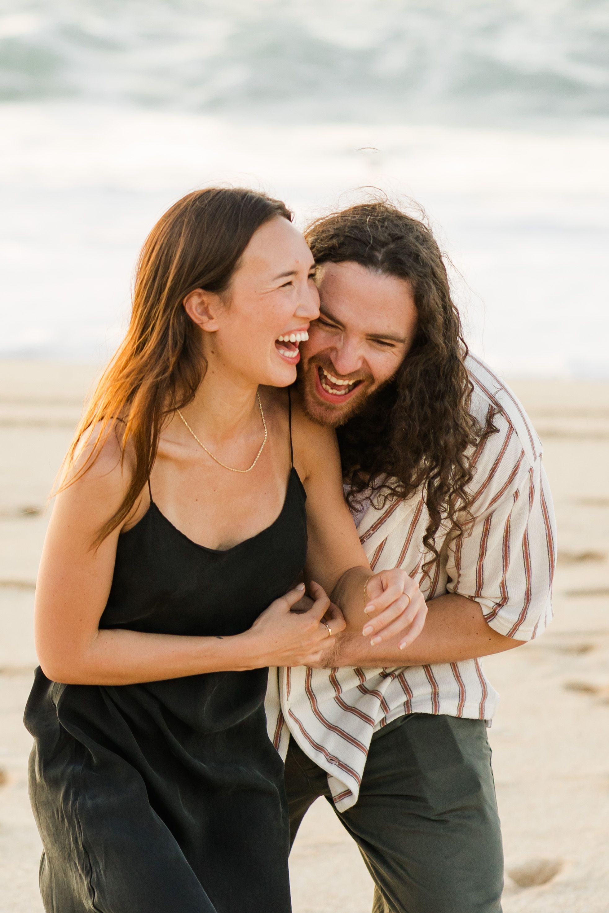 A joyful couple laughs together on a sunny beach during golden hour, sharing a candid moment by the ocean.