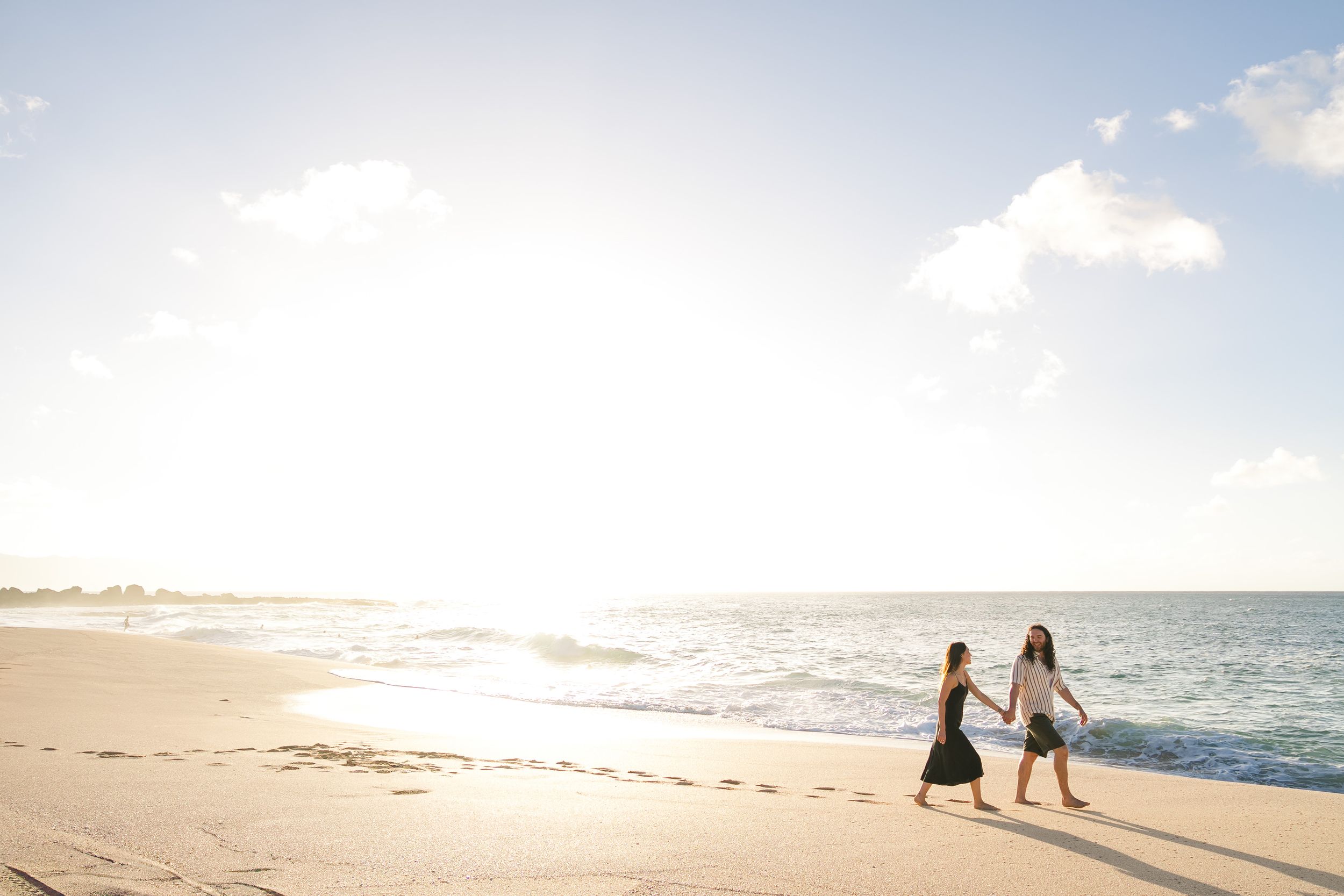 Two people holding hands while walking on a sunny beach at sunset with waves crashing on the shore.