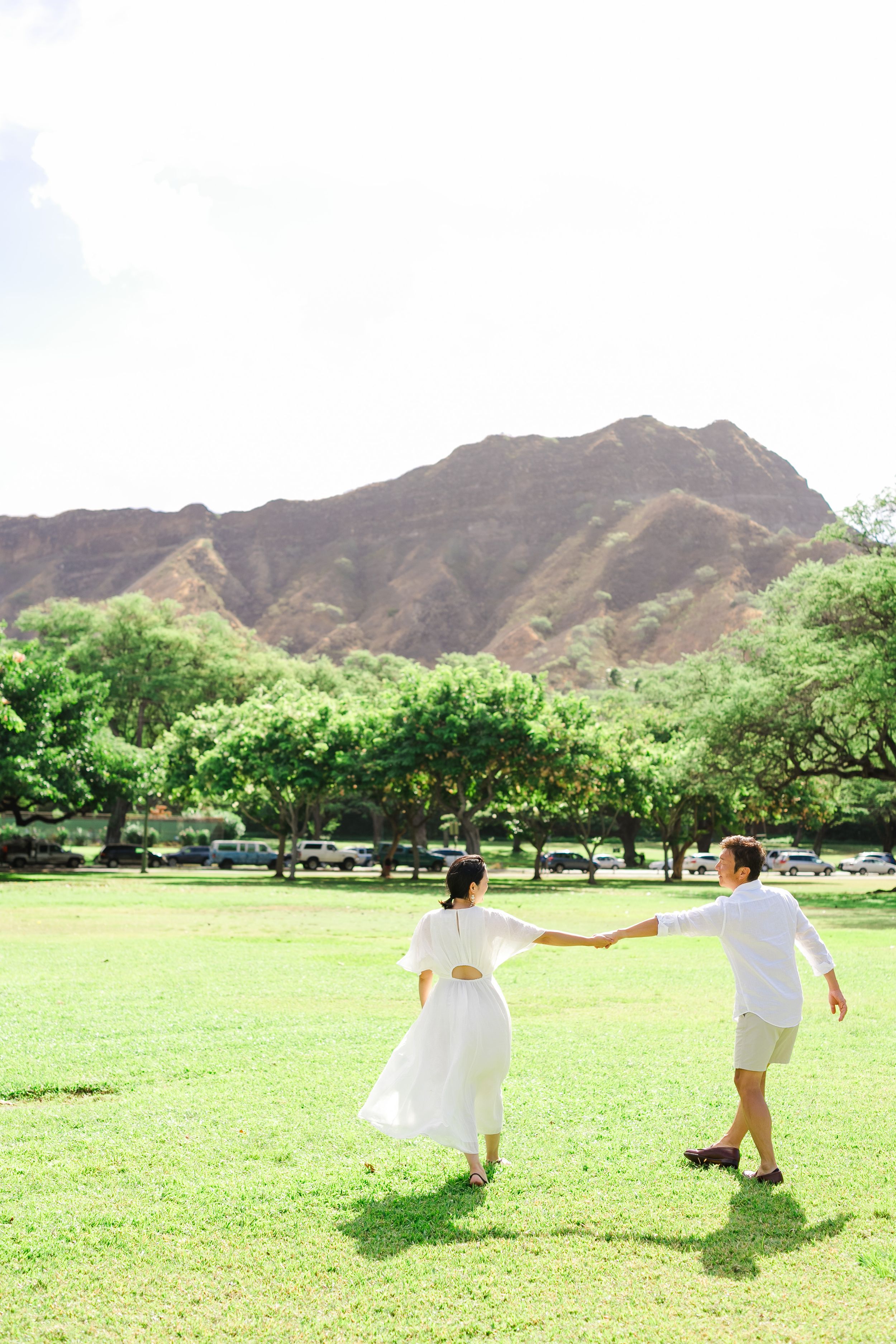 Couple holding hands while walking on grassy field with Diamond Head mountain in background.