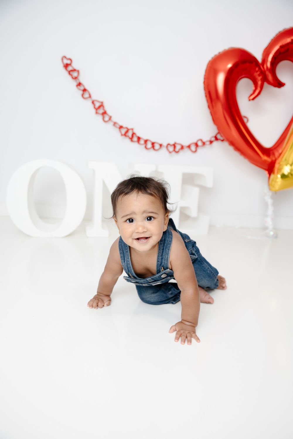 Baby in denim overalls crawls on white backdrop with red heart-shaped balloons and decorative elements.