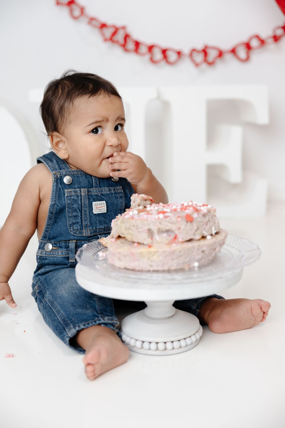 A small child in denim overalls sits beside a decorated pink cake on a white pedestal cake stand.