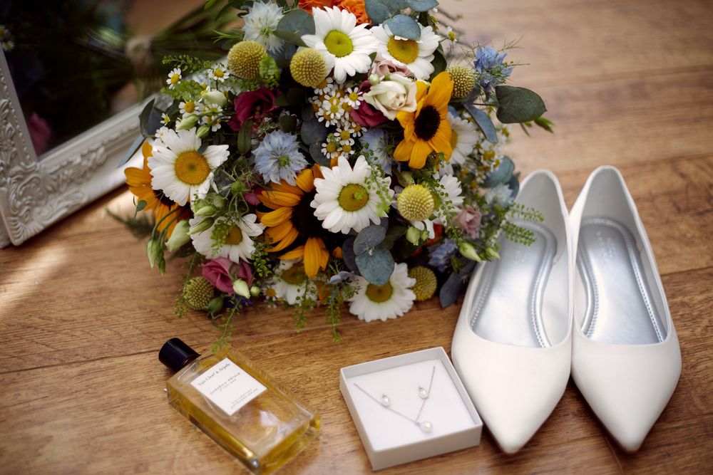 Elegant white bridal shoes and floral bouquets with daisies and sunflowers arranged on wooden surface.