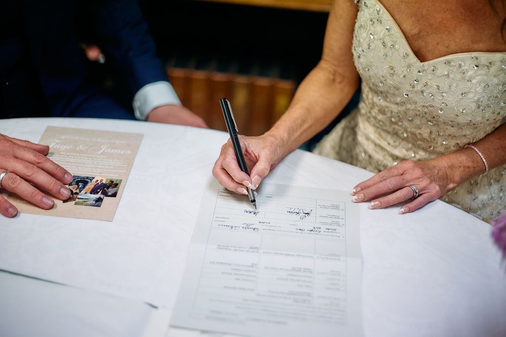 Hands signing legal marriage documents and paperwork with a white wedding dress visible.