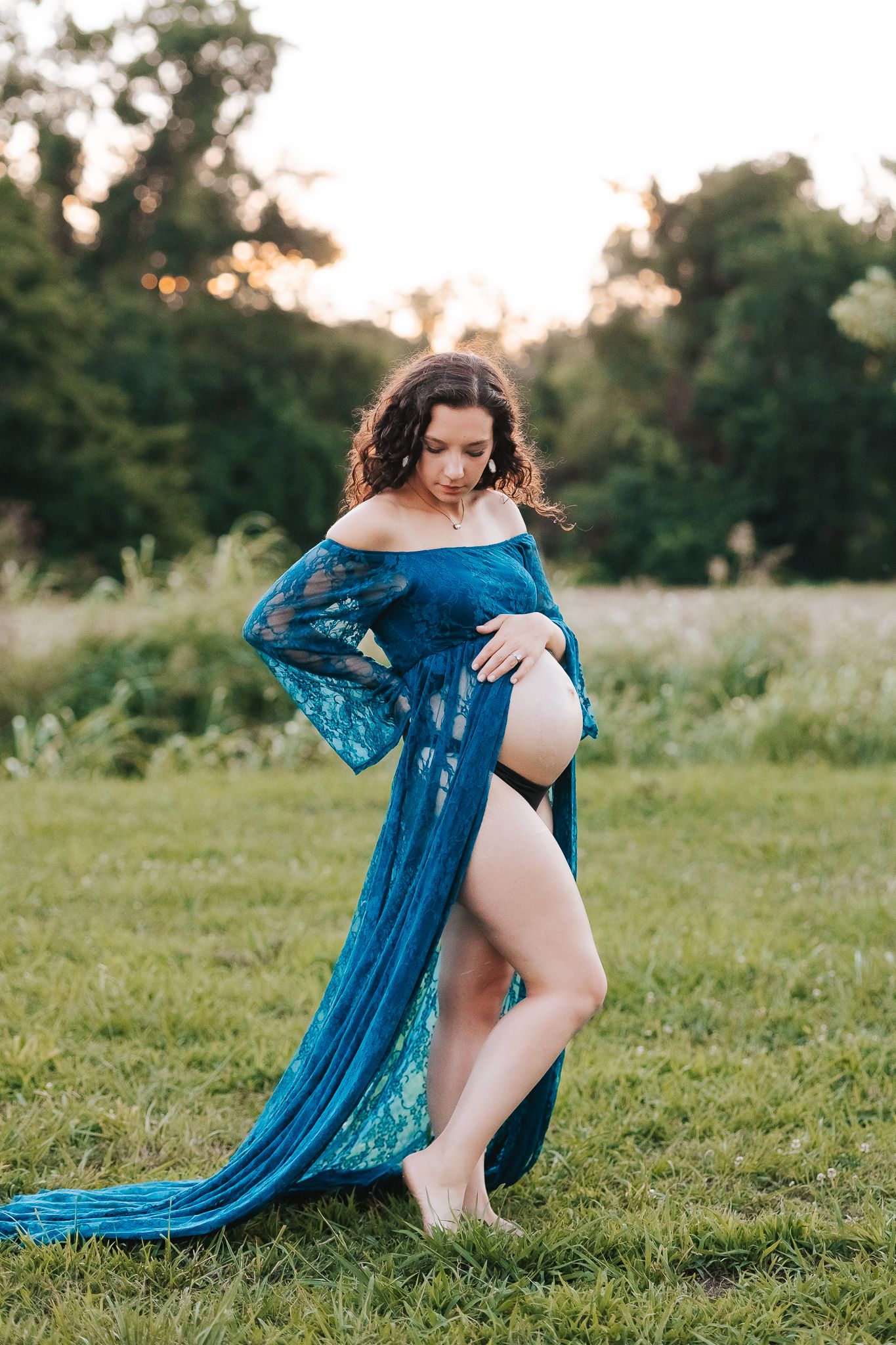 Maternity photo of woman in flowing blue lace dress posing in a field at sunset.