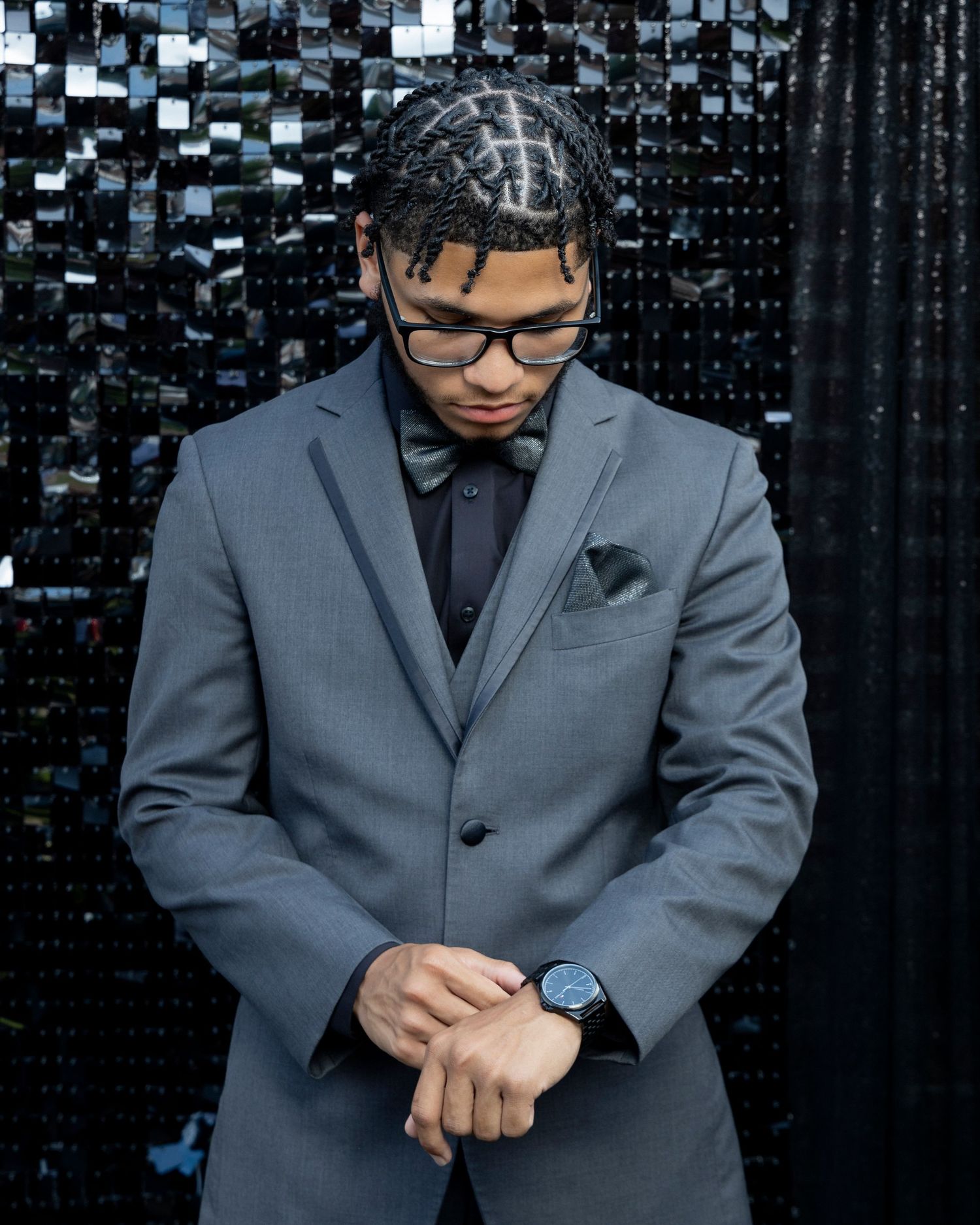 A formal portrait of someone wearing a gray suit and glasses against a dark sparkly background.
