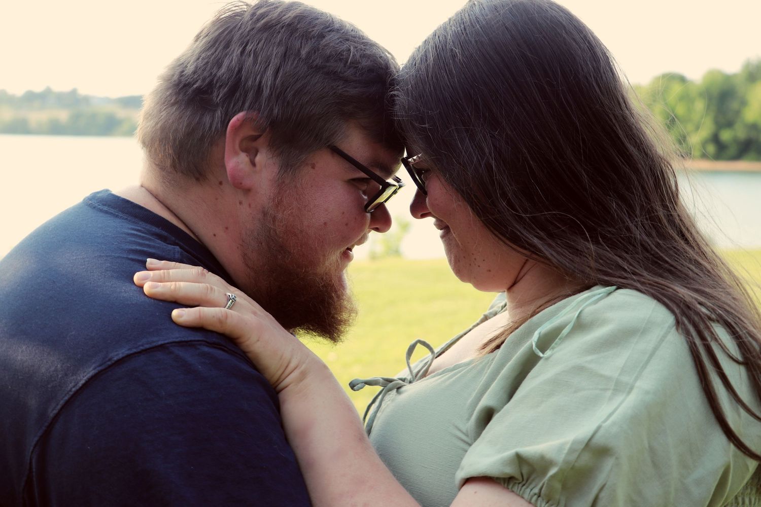 A romantic couple leans their foreheads together near a serene lake in a tender engagement moment.